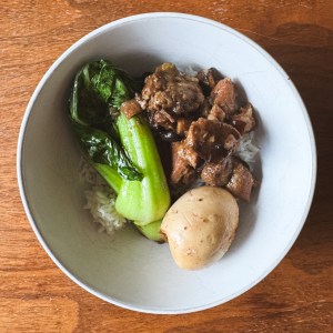 white bowl on a wood table filled with white rice, braised pork, hardboiled egg, and blanched bok choy. 